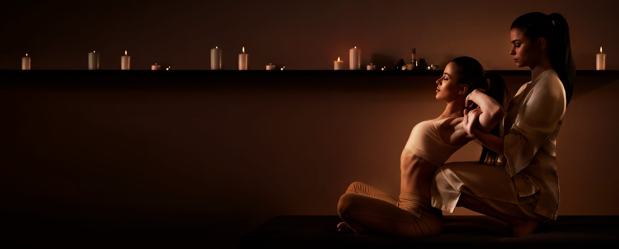 Two women in spa setting with lit candles on shelf behind them