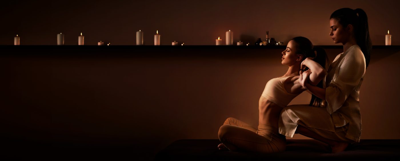 Two women sitting together with candles lit on a shelf behind them in warm lighting